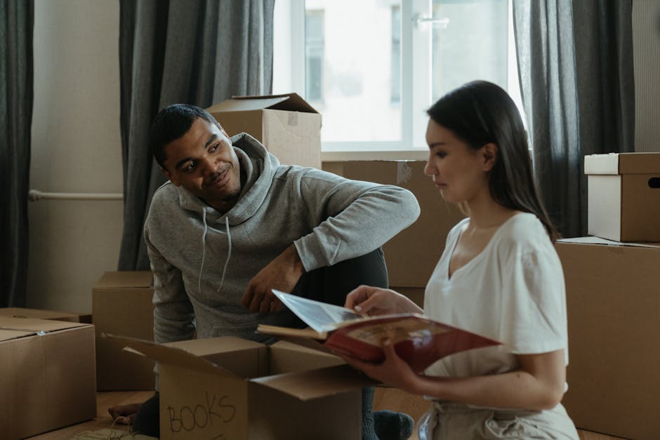 Inside a residential property during a house removal process, a woman with dark hair and a white shirt is seated on the floor, handling packing materials. Behind her, multiple cardboard boxes of vario